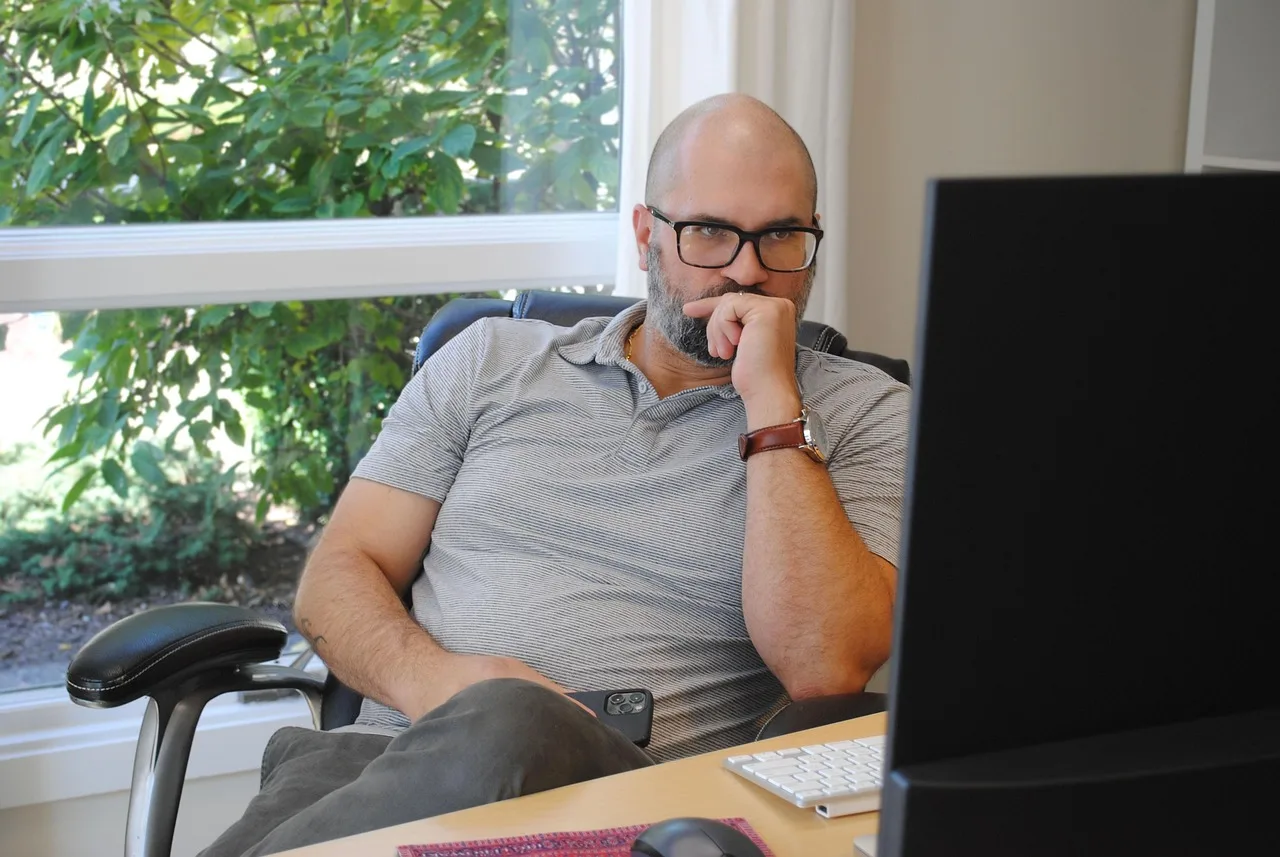 Mężczyzna w okularach siedzi przy biurku i zamyślony patrzy w monitor — ilustracja prokrastynacji